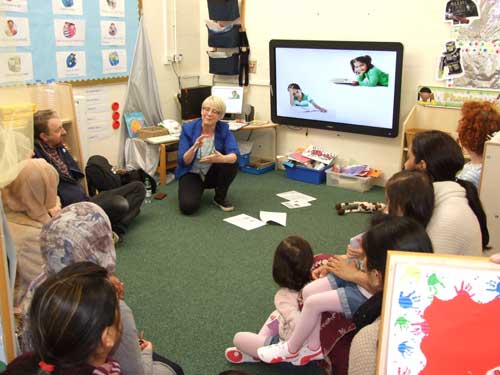 Group of parents and children sitting on the floor in a classroom looking at a screen. 